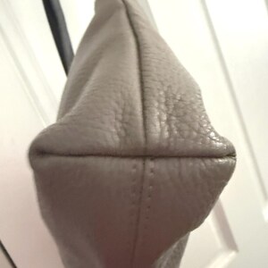May include: A pale grey leather shoulder bag with a silver-tone metal ring and a black strap. The bag has a textured surface and a rounded bottom with stitched seams.