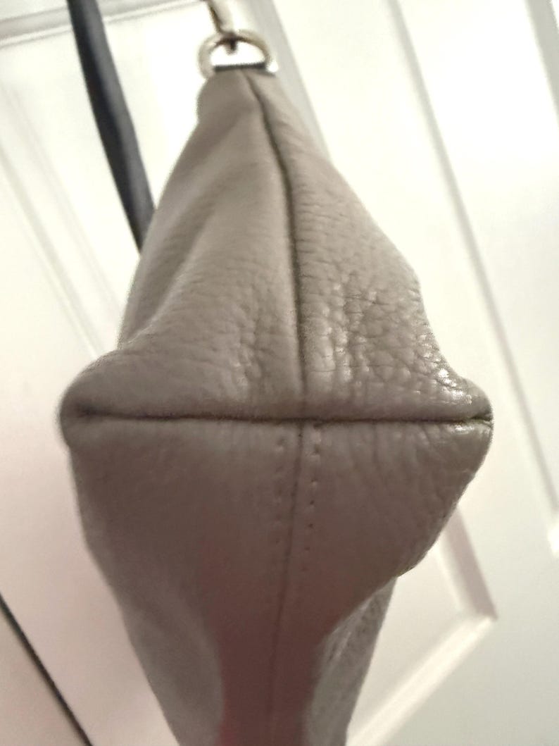 May include: A pale grey leather shoulder bag with a silver-tone metal ring and a black strap. The bag has a textured surface and a rounded bottom with stitched seams.