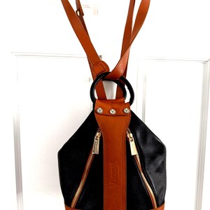 May include: Black and brown leather backpack with gold zippers and hardware. The backpack has a rounded shape with a brown leather strap down the center and brown leather straps for the shoulders. The brand name "Valentina" is visible on the strap.