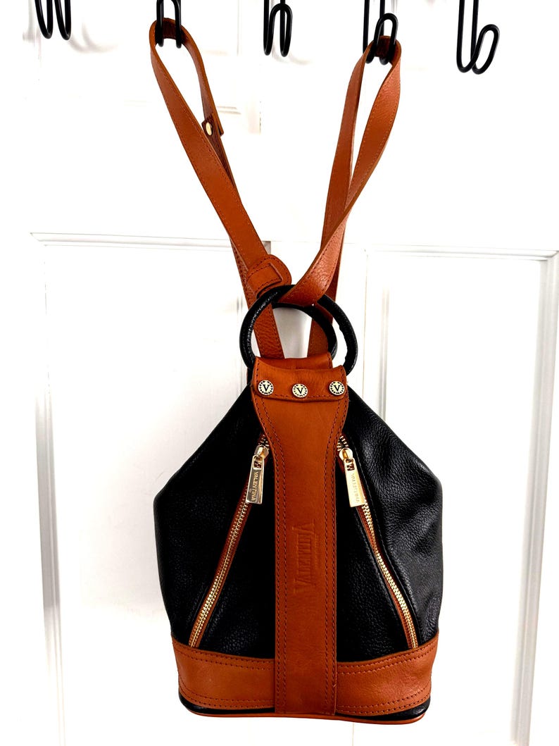 May include: Black and brown leather backpack with gold zippers and hardware. The backpack has a rounded shape with a brown leather strap down the center and brown leather straps for the shoulders. The brand name "Valentina" is visible on the strap.