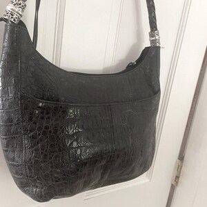 Puede incluir: Bolso de hombro negro con un patr&oacute;n texturizado similar al de un cocodrilo. El bolso tiene una forma curva, una correa trenzada y herrajes decorativos plateados en las fijaciones de la correa. Un bolsillo frontal es visible.