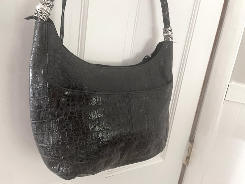Puede incluir: Bolso de hombro negro con un patr&oacute;n texturizado similar al de un cocodrilo. El bolso tiene una forma curva, una correa trenzada y herrajes decorativos plateados en las fijaciones de la correa. Un bolsillo frontal es visible.