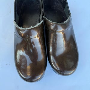 May include: A pair of dark brown clogs with a glossy, patent-like finish. The shoes feature a rounded toe, a high-cut vamp, and a black interior lining with a visible grey trim along the collar edge. The surface of the clogs is smooth.