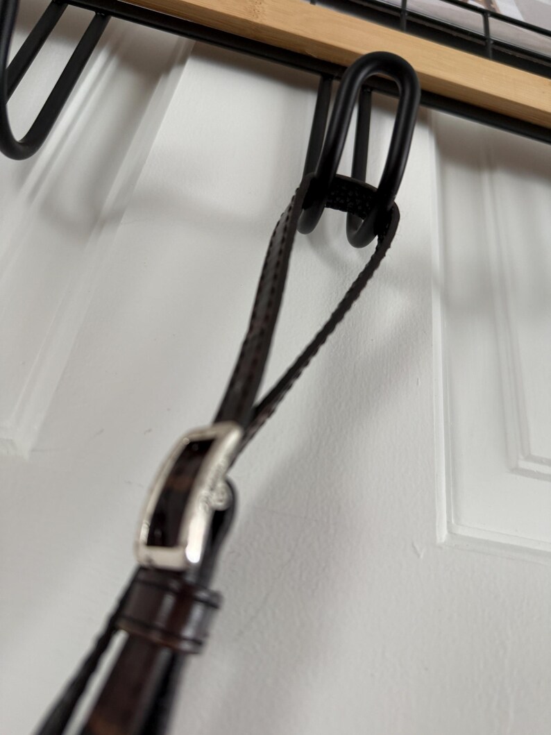 May include: Close-up of a dark brown leather strap with a silver buckle. The strap is attached to a black metal hook, likely for hanging. The background is a white door.