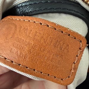 May include: Close-up of a brown leather tag with the embossed text "VALENTINA GENUINE LEATHER CUIR VERITABLE". The tag is stitched with brown thread and is attached to a black leather bag with a gold zipper.