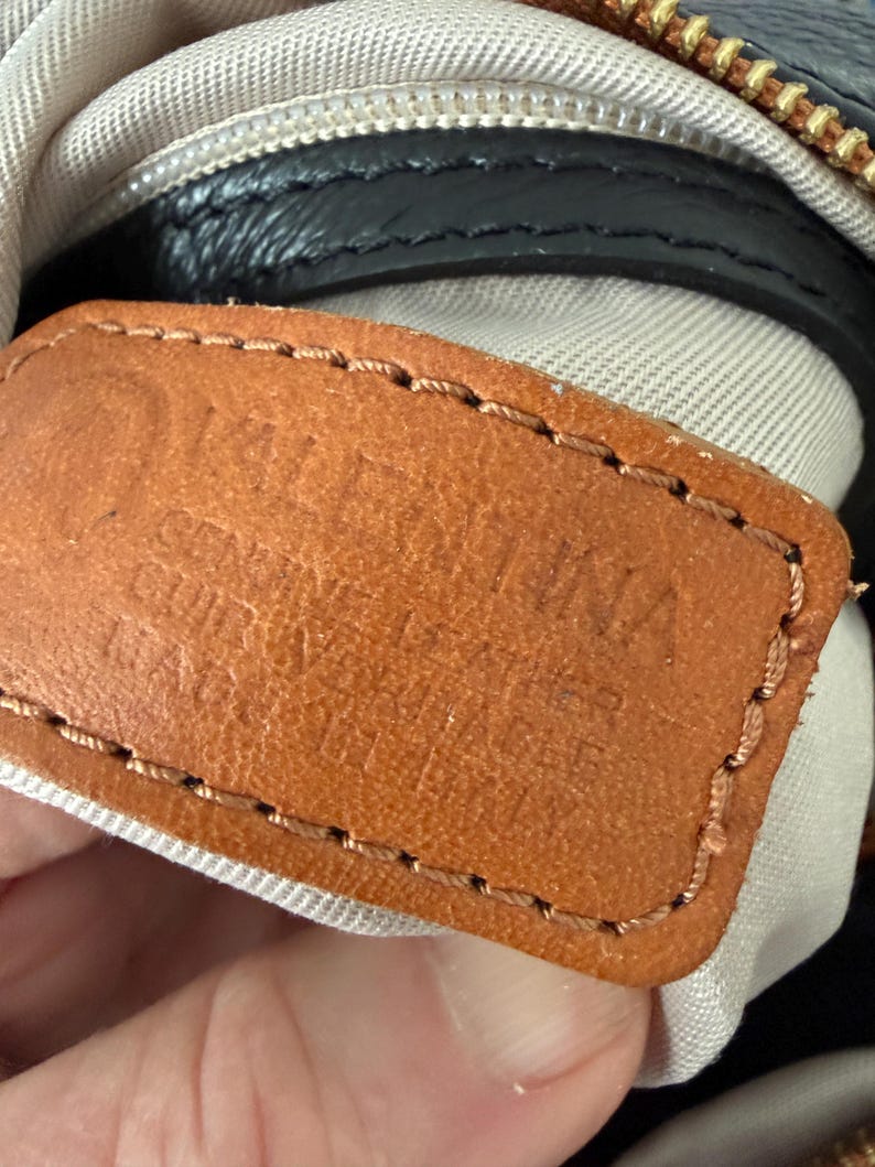 May include: Close-up of a brown leather tag with the embossed text "VALENTINA GENUINE LEATHER CUIR VERITABLE". The tag is stitched with brown thread and is attached to a black leather bag with a gold zipper.