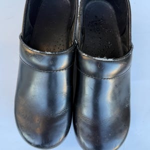 May include: A pair of black leather clogs with a rounded toe, a smooth finish, and a slip-on design. The interior features a dark lining with a perforated footbed, and the exterior shows a clean, solid black surface with visible stitching.