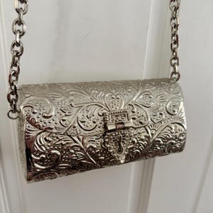 May include: A silver-toned metal clutch bag with an intricate embossed floral and scroll pattern across the entire surface. The rectangular bag features a central hinged clasp closure and is attached to a thick silver-toned metal chain strap.