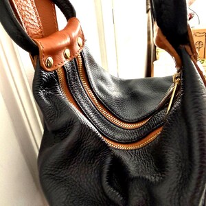 May include: Black leather hobo bag with brown leather accents and gold-tone zippers. The bag features a textured exterior, a curved shape, and a brown leather handle with gold studs. The zippers are a prominent design element.