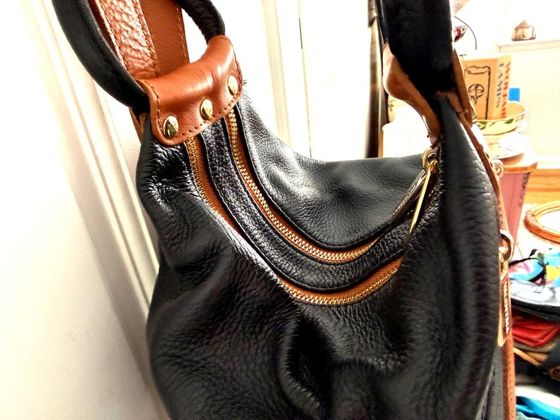 May include: Black leather hobo bag with brown leather accents and gold-tone zippers. The bag features a textured exterior, a curved shape, and a brown leather handle with gold studs. The zippers are a prominent design element.