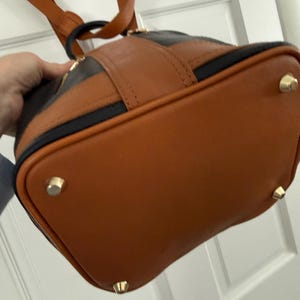 May include: A brown and black leather backpack with gold-colored feet. The bag has a rounded shape with a brown base and a black stripe across the top. The straps are brown.