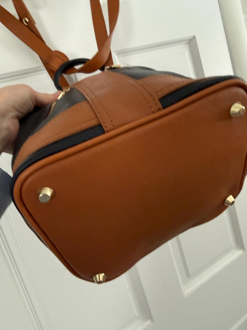 May include: A brown and black leather backpack with gold-colored feet. The bag has a rounded shape with a brown base and a black stripe across the top. The straps are brown.