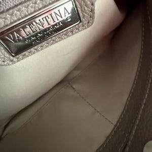 May include: A close-up of a taupe leather handbag's interior. The bag features a silver metal plate with the name "VALENTINA" embossed on it. The interior lining is a light beige colour, and the bag has visible stitching.