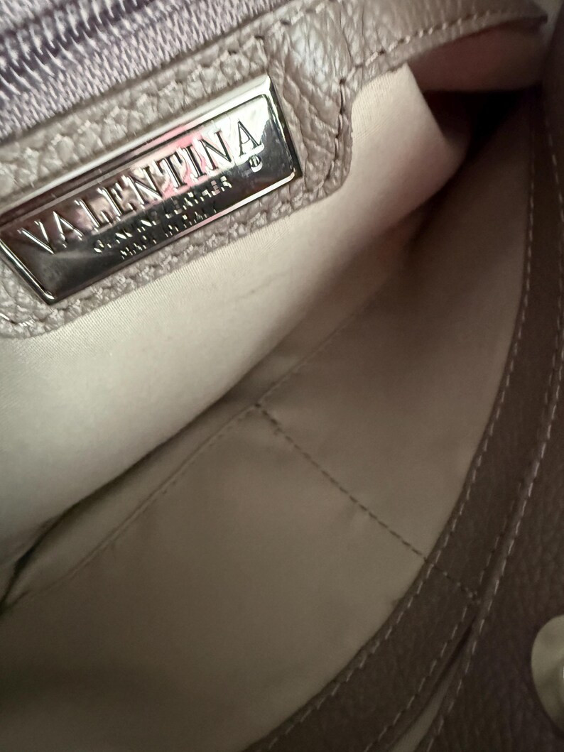 May include: A close-up of a taupe leather handbag's interior. The bag features a silver metal plate with the name "VALENTINA" embossed on it. The interior lining is a light beige colour, and the bag has visible stitching.