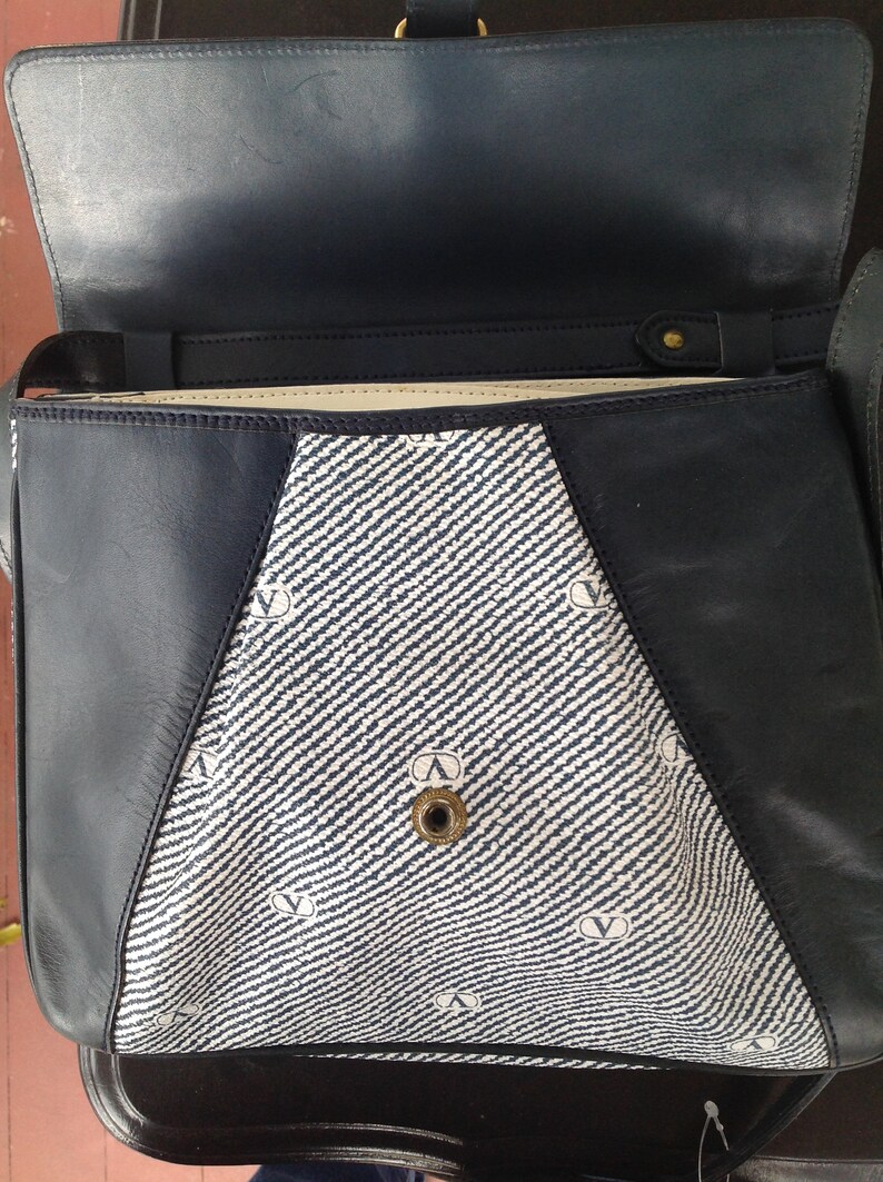 May include: A blue and white striped fabric handbag with a black leather trim. The bag has a flap closure and a gold metal buckle. The bag is lined with a cream-colored fabric.