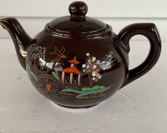 May include: A brown ceramic teapot with a lid, featuring a hand-painted scene of a pagoda, bridge, and trees in gold, red, green, and blue.