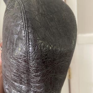 Puede incluir: Primer plano de un bolso de cuero negro con un patr&oacute;n texturizado similar al de un cocodrilo. El bolso tiene una forma curva y una costura visible en el lateral. El cuero parece desgastado y tiene un ligero brillo.