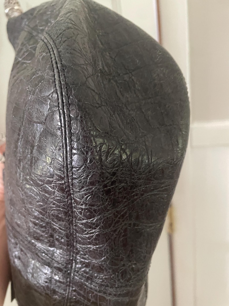 Puede incluir: Primer plano de un bolso de cuero negro con un patr&oacute;n texturizado similar al de un cocodrilo. El bolso tiene una forma curva y una costura visible en el lateral. El cuero parece desgastado y tiene un ligero brillo.