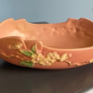 May include: A pink ceramic bowl with a floral design. The bowl has two handles that resemble branches. The floral design features pink and yellow flowers and green leaves.
