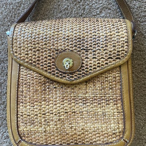 May include: A woven crossbody bag with a flap closure. The bag features a light brown woven pattern with tan leather trim and a matching shoulder strap. The flap has a gold-colored clasp. The bag is rectangular in shape.