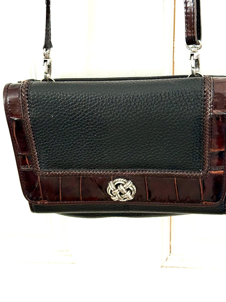 May include: Black and brown leather crossbody bag with a silver-tone clasp. The bag features a textured black leather center panel, framed by brown, crocodile-embossed leather. A silver-tone chain strap is attached.
