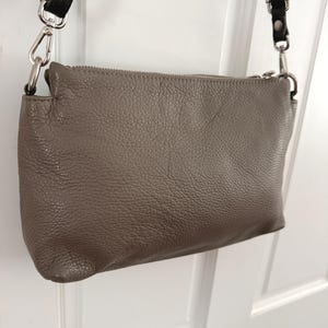 May include: A taupe-coloured crossbody bag with a textured leather exterior. The bag features a silver zip and silver metal hardware. It has a black leather strap with silver clasps for attachment.