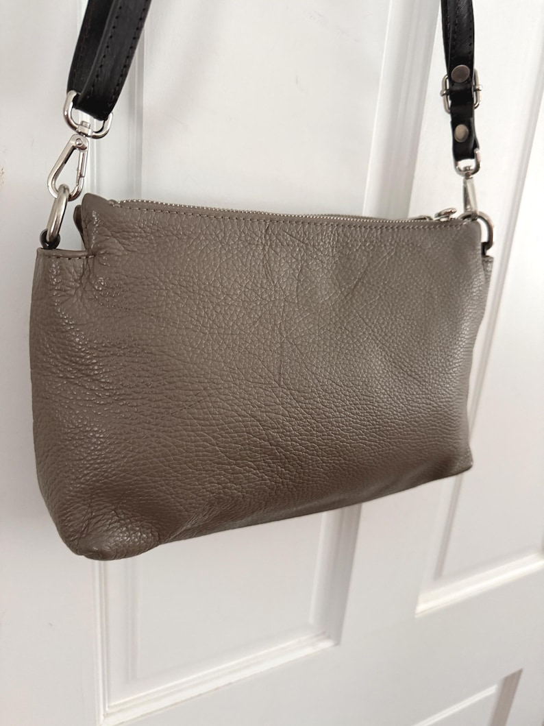 May include: A taupe-coloured crossbody bag with a textured leather exterior. The bag features a silver zip and silver metal hardware. It has a black leather strap with silver clasps for attachment.
