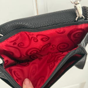 May include: Black leather crossbody bag with a red patterned interior. The bag has a silver-tone zipper and hardware, and a brown braided strap. The interior lining features a swirling design.