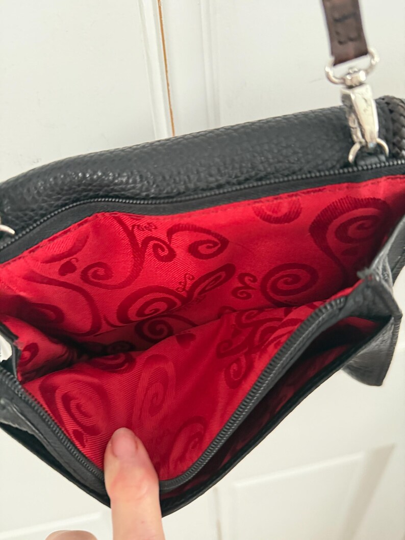 May include: Black leather crossbody bag with a red patterned interior. The bag has a silver-tone zipper and hardware, and a brown braided strap. The interior lining features a swirling design.