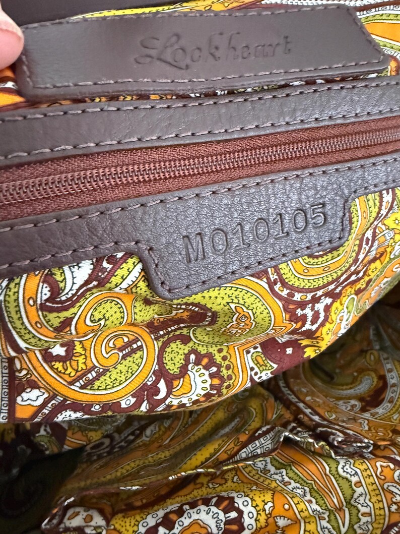 K&ouml;nnte beinhalten: Nahaufnahme des Innenraums einer braunen Lederhandtasche. Die Tasche hat ein Paisley-Musterfutter in Gelb-, Orange- und Braunt&ouml;nen. Der Markenname "Lookheart" ist auf einem Lederetikett eingepr&auml;gt. Die Modellnummer "MO10105" ist ebenfalls sichtbar.