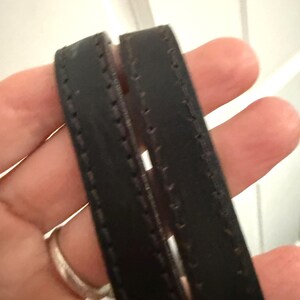 May include: Close-up of two black leather straps with visible stitching. The straps appear to be part of a handbag or tote, with a focus on the texture and craftsmanship of the material.