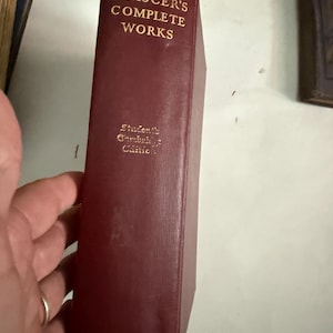May include: A burgundy-colored book titled "Chaucer's Complete Works" with gold lettering. The book is a Student's Cambridge Edition, published by Houghton Mifflin Co. The book is held by a hand.