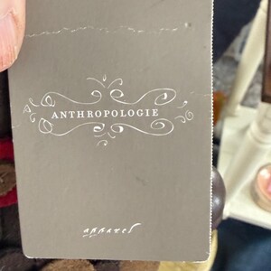 K&ouml;nnte beinhalten: Ein graues Etikett mit dem Wort "ANTHROPOLOGIE" in wei&szlig;er Schrift. Das Etikett ist an einem Kleidungsst&uuml;ck befestigt. Das Wort "apparel" ist in kleinerer Schrift am unteren Rand des Etiketts aufgedruckt.