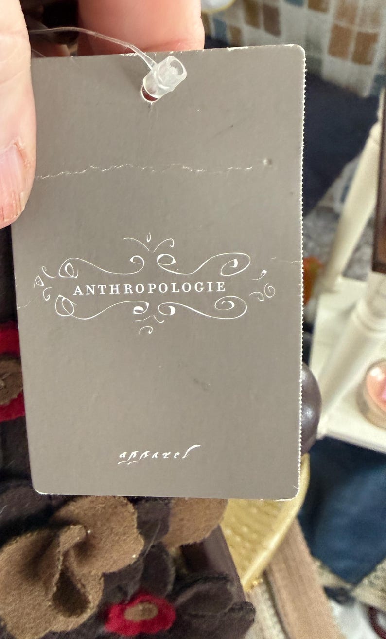 K&ouml;nnte beinhalten: Ein graues Etikett mit dem Wort "ANTHROPOLOGIE" in wei&szlig;er Schrift. Das Etikett ist an einem Kleidungsst&uuml;ck befestigt. Das Wort "apparel" ist in kleinerer Schrift am unteren Rand des Etiketts aufgedruckt.