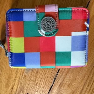 May include: A colorful wallet with a patchwork design in shades of blue, green, red, orange, and yellow. The wallet has a silver-tone button closure and a zipper. The brand logo is visible on the button.