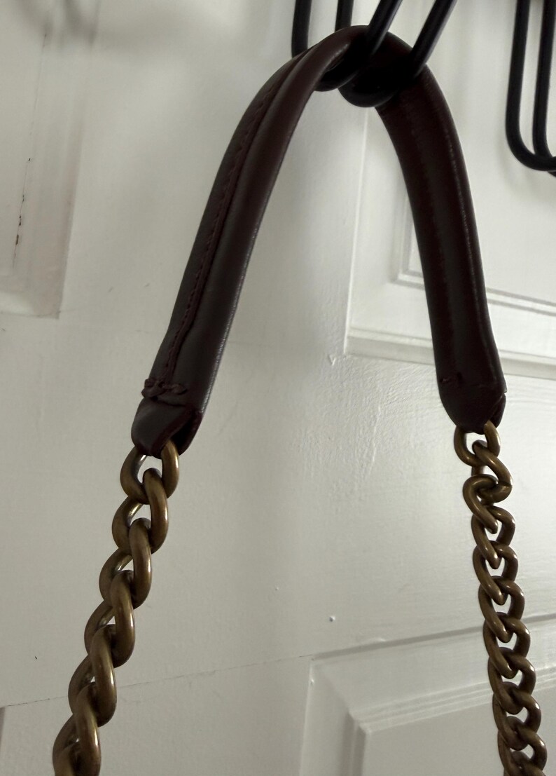 K&ouml;nnte beinhalten: Nahaufnahme eines burgunderfarbenen Lederhandtaschengriffs mit goldfarbenen Kettentr&auml;gern. Der Griff ist an einem schwarzen Metallhaken befestigt. Der Hintergrund ist eine wei&szlig;e T&uuml;r.
