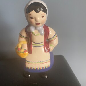 May include: A ceramic figurine of a girl wearing a blue and white headscarf, a red vest, and a white dress with colorful trim. She is holding a yellow basket with red and yellow flowers.