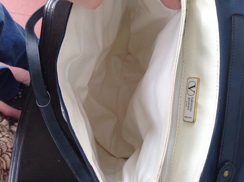 May include: The inside of a navy blue handbag with a white lining. The bag has a gold metal label that reads "valentino garavani made in italy".