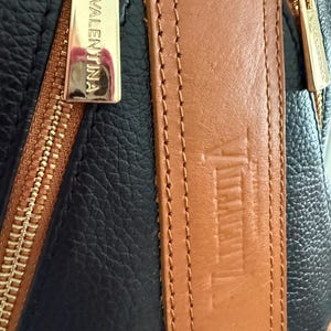 May include: Close-up of a black and brown leather bag with gold-colored zippers. The brand name "VALENTINA" is visible on the zipper pull and strap. The bag has a textured leather appearance.