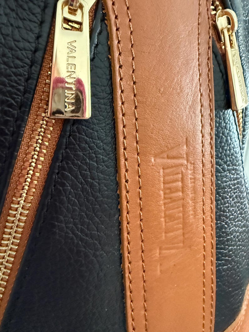 May include: Close-up of a black and brown leather bag with gold-colored zippers. The brand name "VALENTINA" is visible on the zipper pull and strap. The bag has a textured leather appearance.