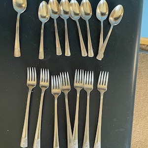 May include: A set of 12 silver-plated teaspoons and 12 silver-plated forks. The spoons and forks have an ornate design and are arranged in two rows on a black surface.