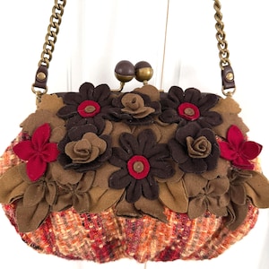 K&ouml;nnte beinhalten: Eine Clutch-Tasche mit Kette, verziert mit Filzblumen. Der Taschenkorpus besteht aus einem tweedartigen Stoff in Orange, Rot und Braun. Braune, rote und beige Filzblumen sind auf der Oberseite angeordnet und bilden ein strukturiertes Design.