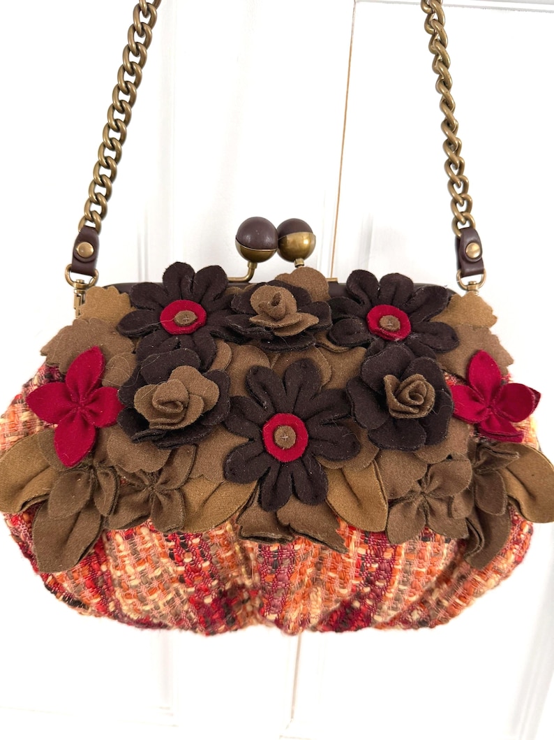 K&ouml;nnte beinhalten: Eine Clutch-Tasche mit Kette, verziert mit Filzblumen. Der Taschenkorpus besteht aus einem tweedartigen Stoff in Orange, Rot und Braun. Braune, rote und beige Filzblumen sind auf der Oberseite angeordnet und bilden ein strukturiertes Design.