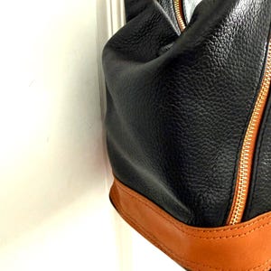 May include: Black leather handbag with a textured finish, featuring a tan leather base and trim. The bag has a gold zipper and brown accents.
