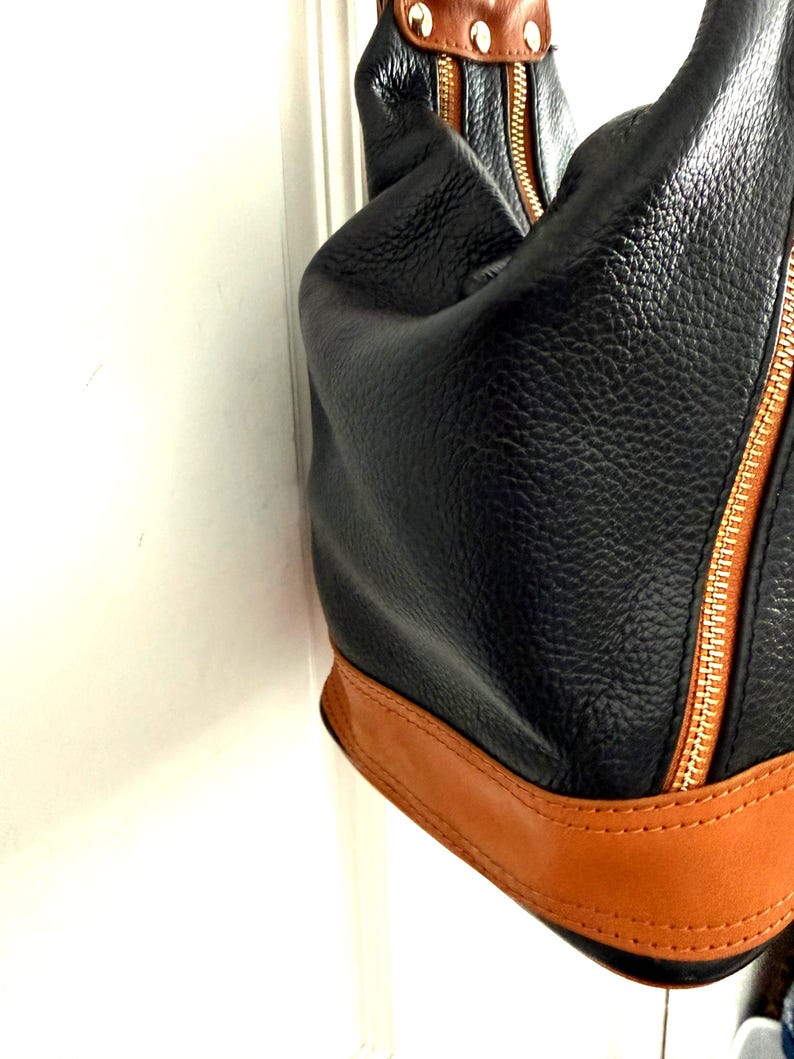 May include: Black leather handbag with a textured finish, featuring a tan leather base and trim. The bag has a gold zipper and brown accents.