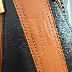 May include: Close-up of a black and brown leather handbag. The bag features a brown leather strap with the embossed word "Valentina". The strap is stitched to the black leather body of the bag with a gold zipper.
