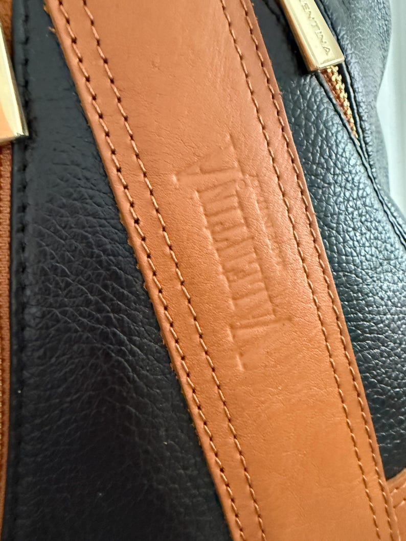 May include: Close-up of a black and brown leather handbag. The bag features a brown leather strap with the embossed word "Valentina". The strap is stitched to the black leather body of the bag with a gold zipper.