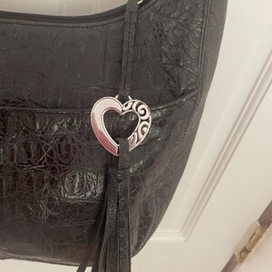 Puede incluir: Bolso de hombro negro con un patr&oacute;n texturizado similar al de un cocodrilo. El bolso presenta un dije plateado en forma de coraz&oacute;n con un dise&ntilde;o decorativo y una borla negra. Tiene cierre de cremallera y un bolsillo peque&ntilde;o.