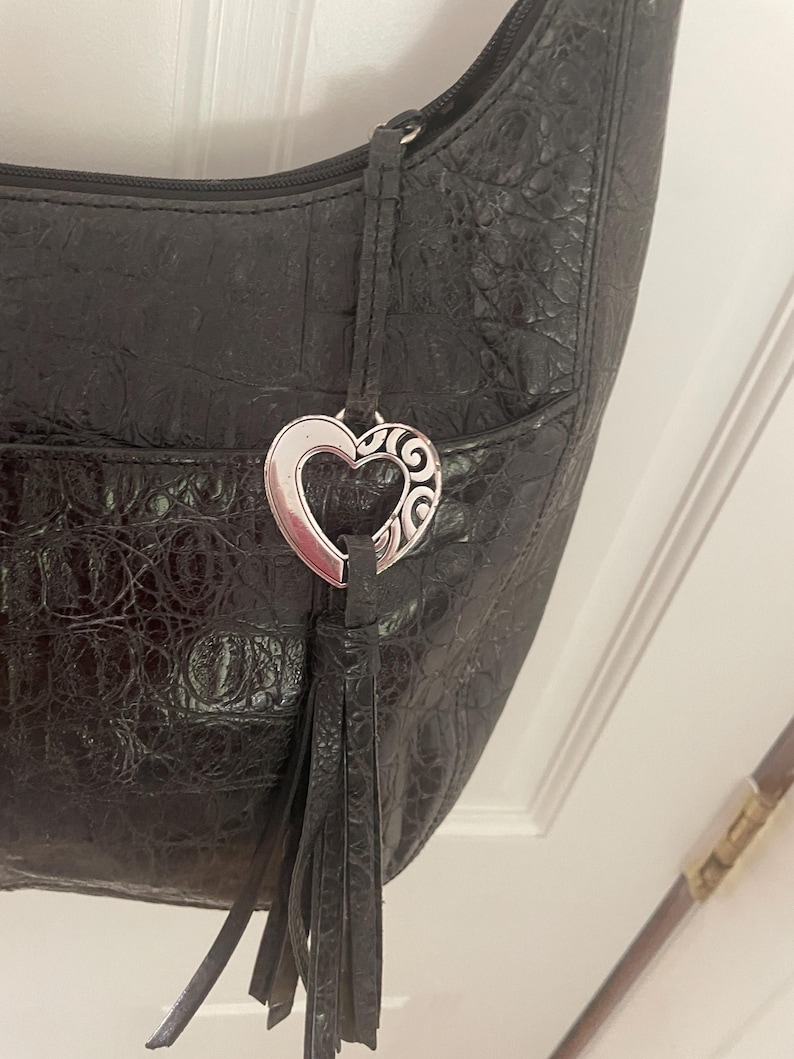 Puede incluir: Bolso de hombro negro con un patr&oacute;n texturizado similar al de un cocodrilo. El bolso presenta un dije plateado en forma de coraz&oacute;n con un dise&ntilde;o decorativo y una borla negra. Tiene cierre de cremallera y un bolsillo peque&ntilde;o.