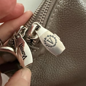 May include: Close-up of a grey leather handbag with a silver zip and zip pulls. The zip pulls have the brand name "Vagrant" engraved on them. The handbag has a textured surface.
